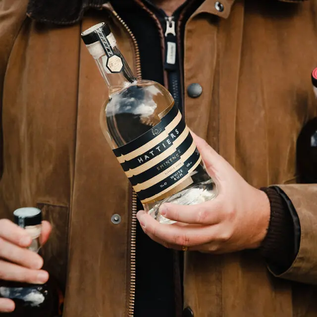 A man in a brown wax jacket holding a Hattiers White rum bottle in his hand
